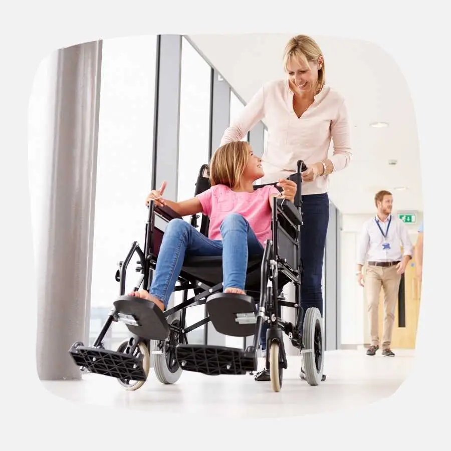 A smiling child in a wheelchair being pushed by a caregiver in a bright hospital corridor.