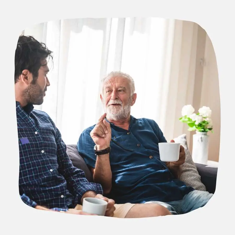An older man with a cup of coffee in hand, engaged in a conversation with a younger man at home.