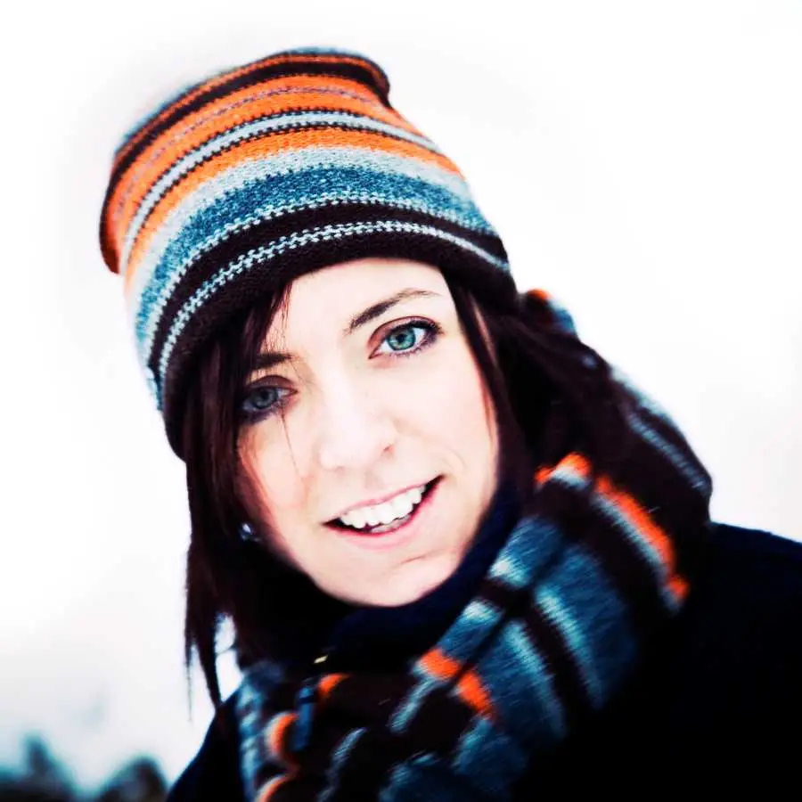 Woman wearing a striped beanie and scarf in a snowy environment, smiling at the camera.