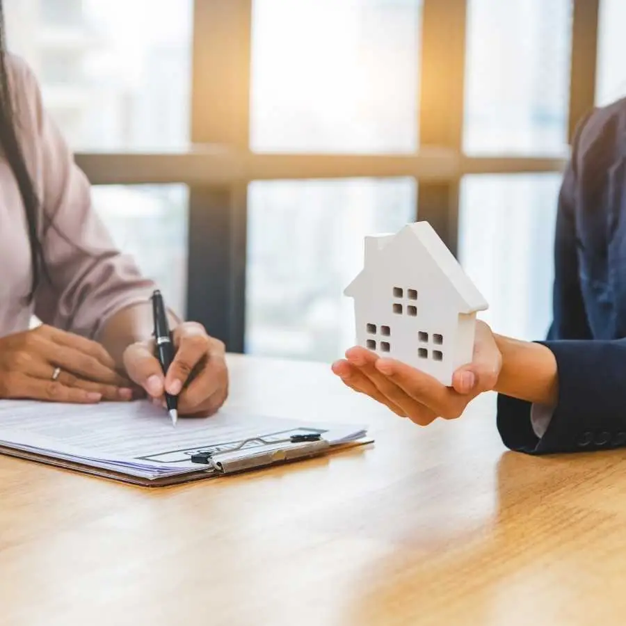 Person holding a small house model while another signs documents